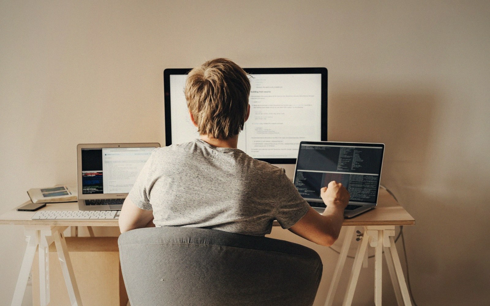 Person seen from behind working at a desk with a monitor and two laptops
