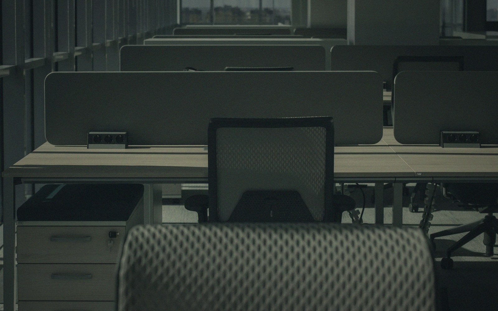 Empty office cubicles with mesh chairs and dividers lit by dim natural light