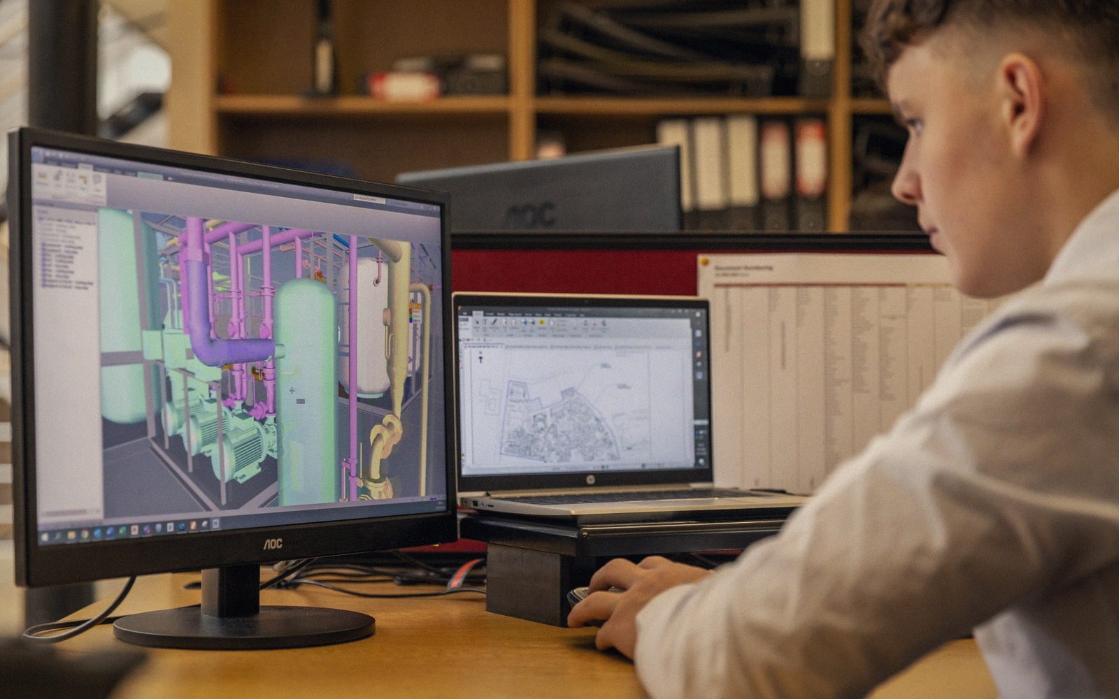 An engineer reviews a 3D piping model on a monitor beside a laptop showing a site plan