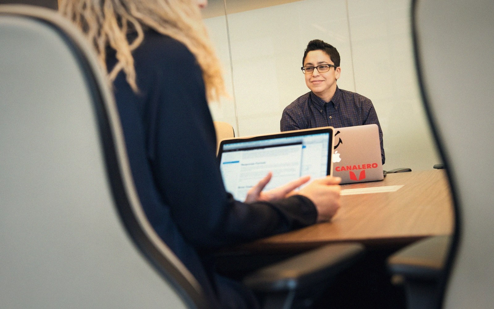 Two coworkers sit at a conference table with open laptops during a meeting