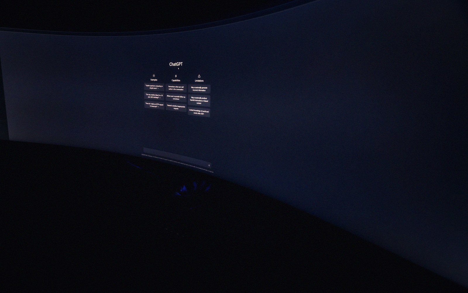 Curved computer monitor displaying the ChatGPT homepage with example prompts in a dark room